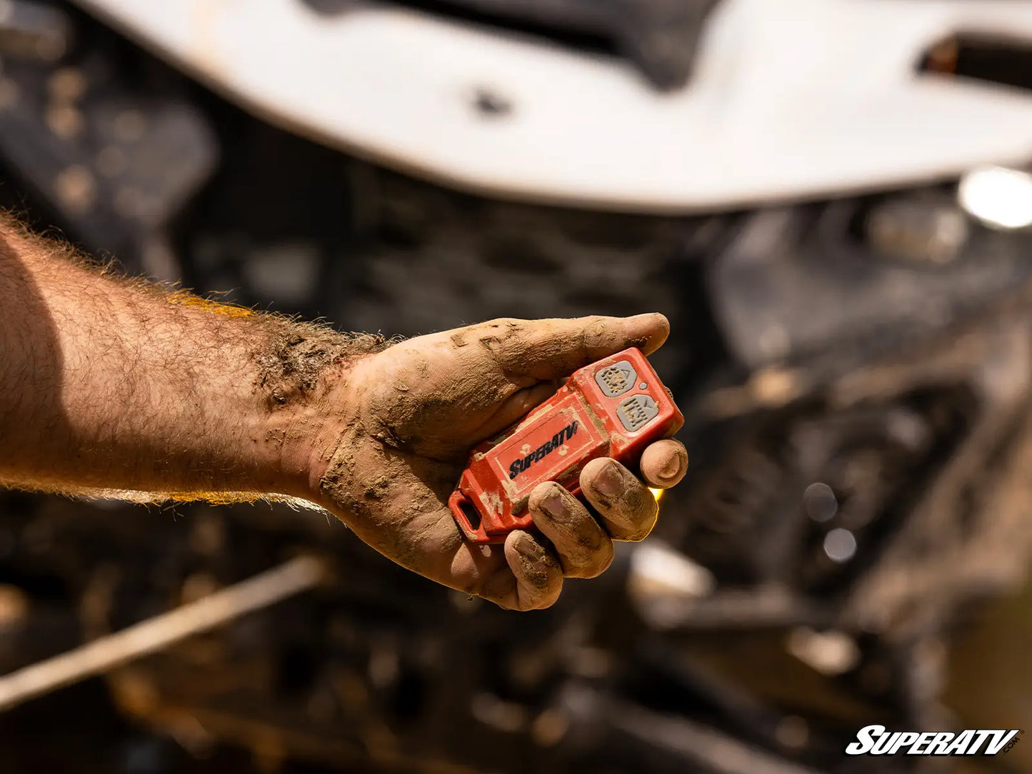 Hand holding a red SuperATV device with a blurred off-road vehicle in the background