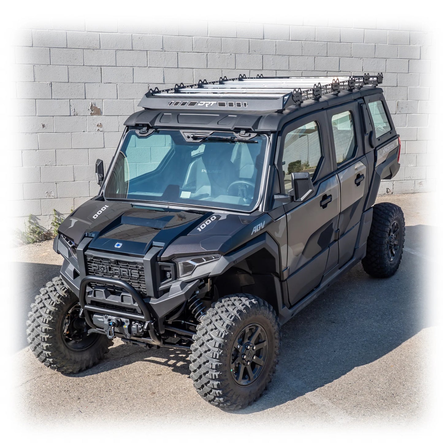 Black off-road vehicle with roof rack against a light gray brick wall.