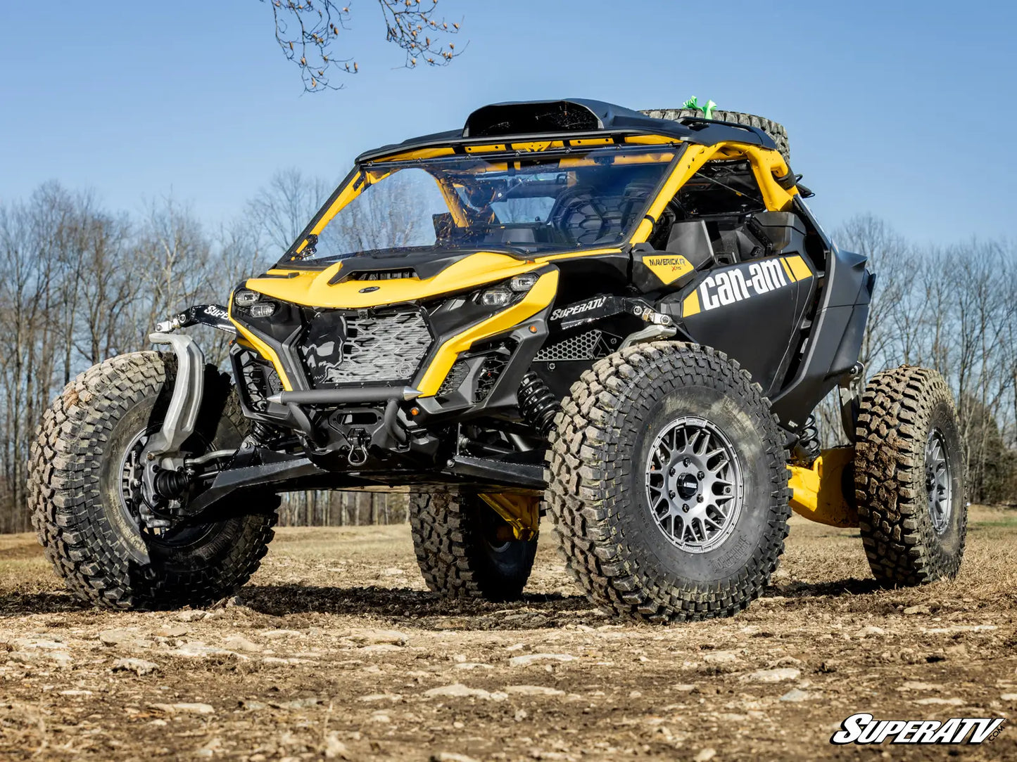 Yellow and black Can-Am off-road vehicle on a dirt field with trees in the background