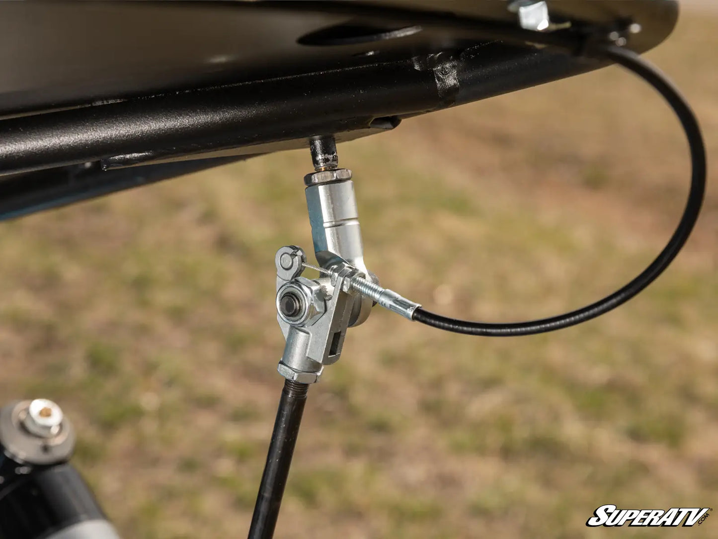 Close-up of a mechanical component on a SuperATV vehicle, with a blurred grassy background.