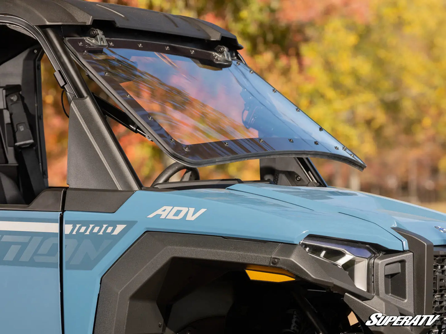 Close-up of a blue SuperATV vehicle with a windshield and front grille, blurred natural background.