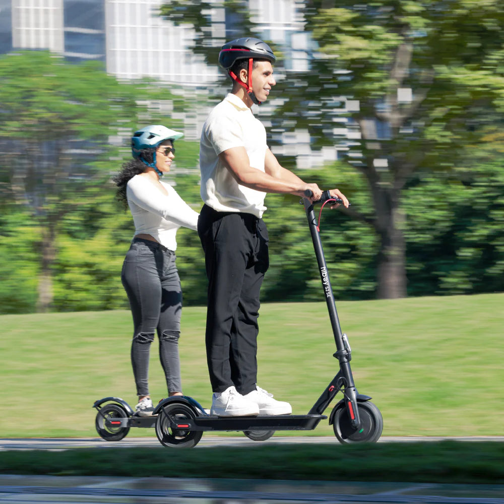 Two people riding electric scooters on a path with trees and buildings in the background