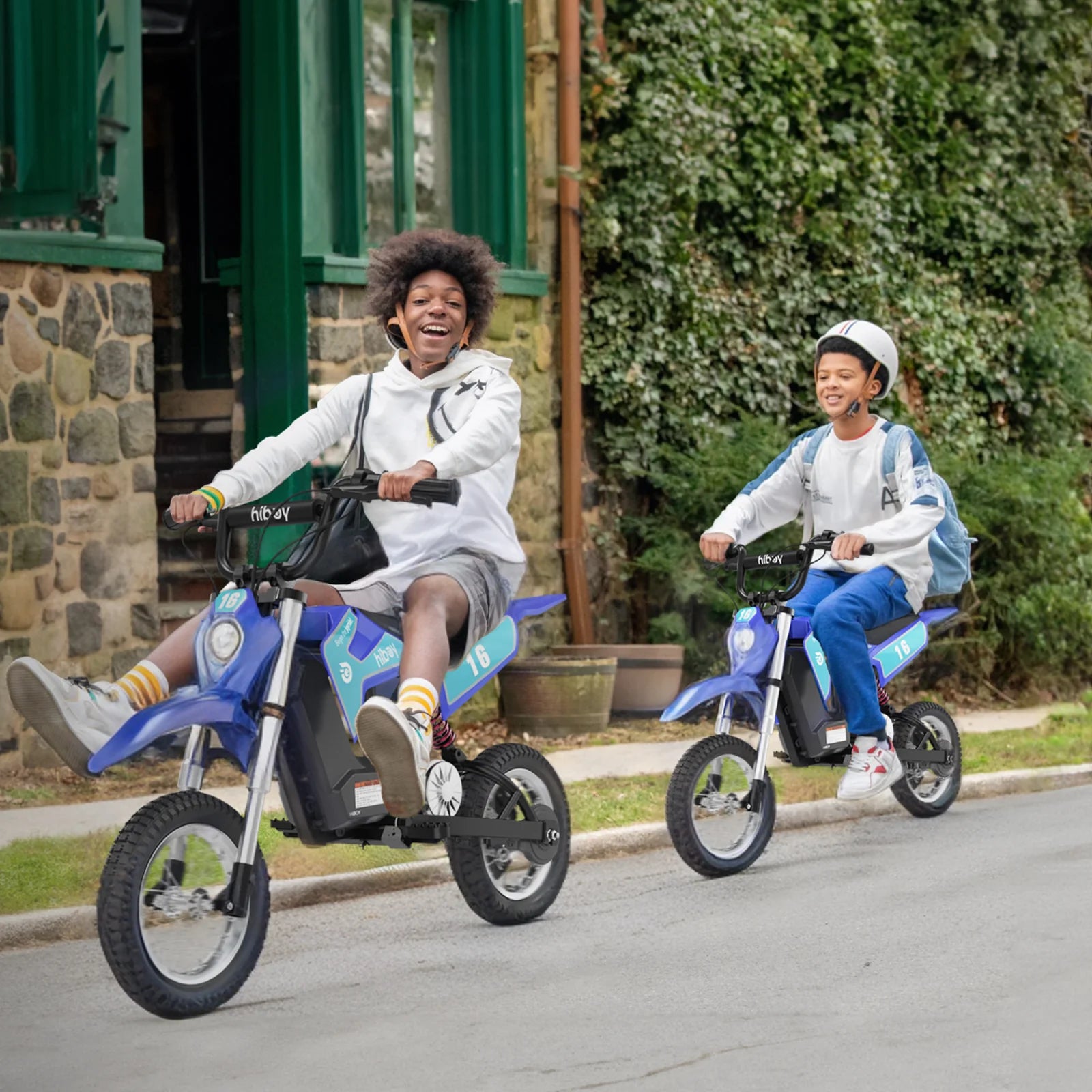 Two children riding small blue motorcycles on a street with a stone building and greenery in the background.