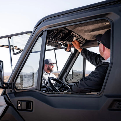 A man in a black cap and black jacket installs or adjusts the windshield sun visor inside a black off-road vehicle, with another man visible in the driver's seat. The setting is outdoors in a desert landscape.