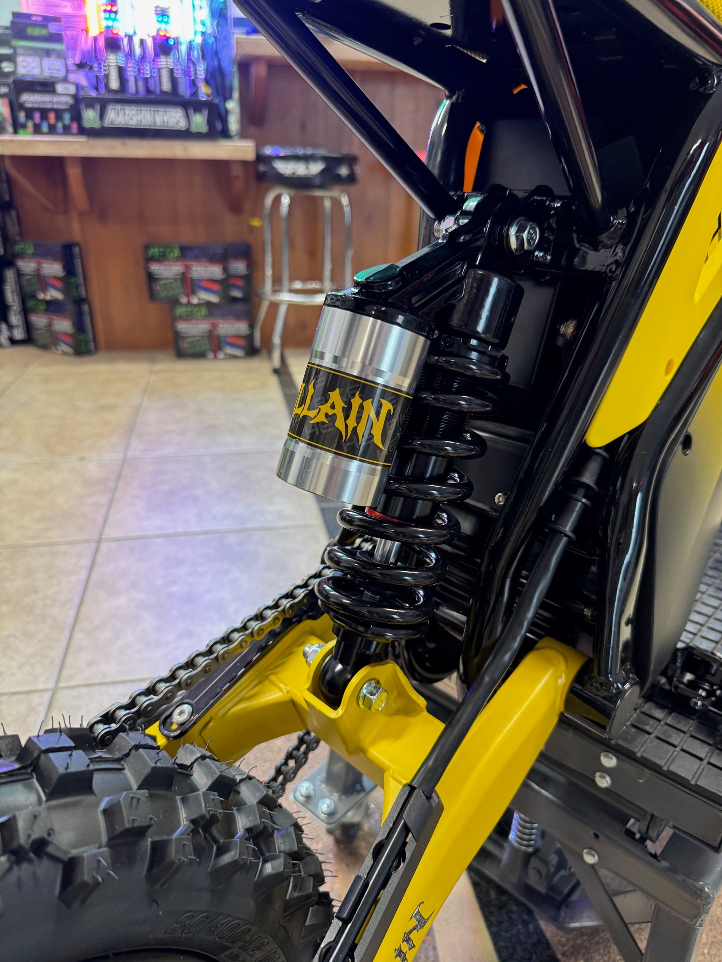Close-up of a motorcycle chain and sprocket with a brand logo on a blurred indoor background.