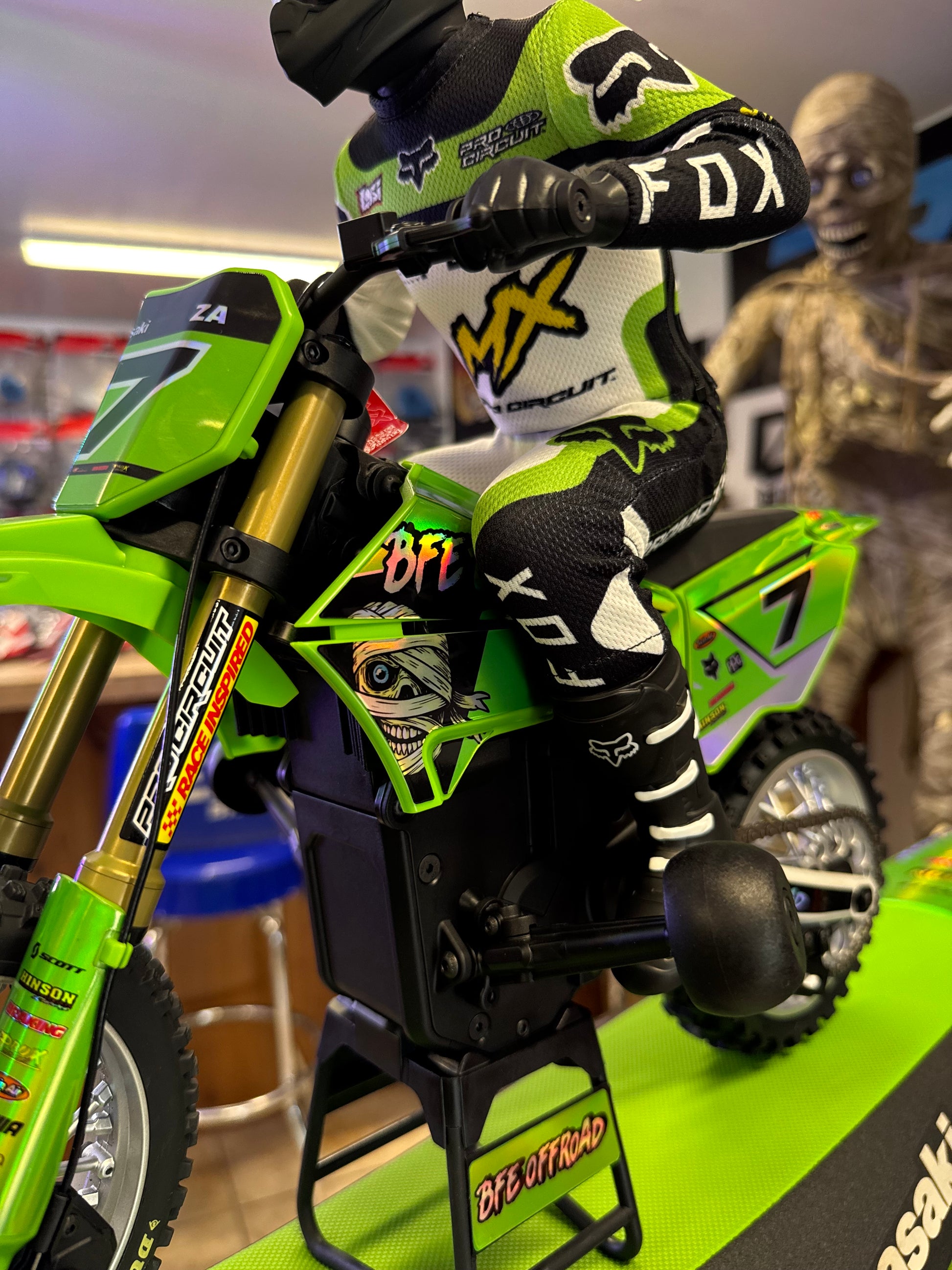 A close-up of a green and black remote control dirt bike with a rider's gloved foot and hand in a racing suit, showing Fox Racing branding on the gloves and bike decals.