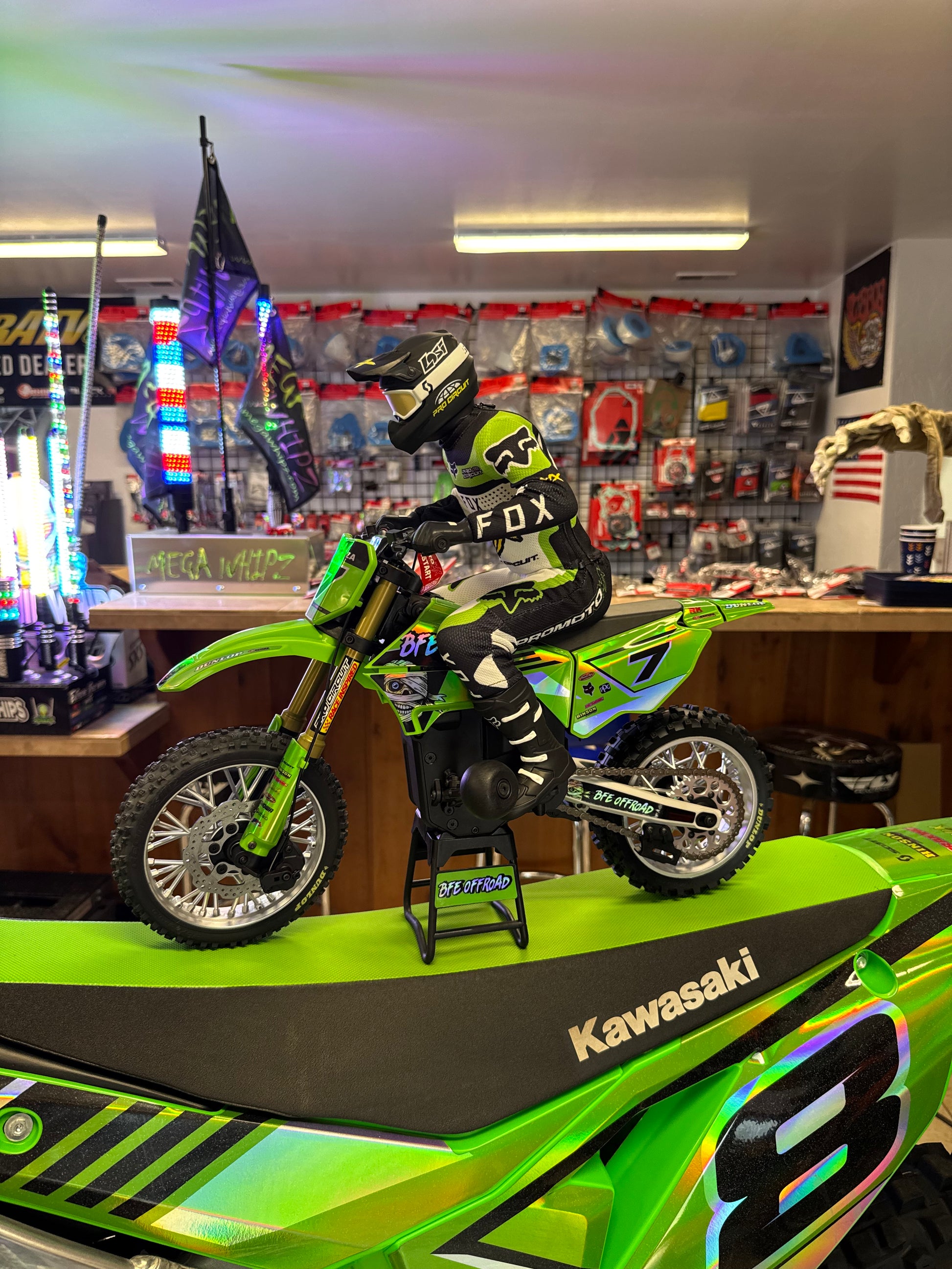 A green and black Kawasaki dirt bike with a rider in Fox racing gear posed on top of it, displayed inside a retail showroom with shelves of motocross merchandise in the background.