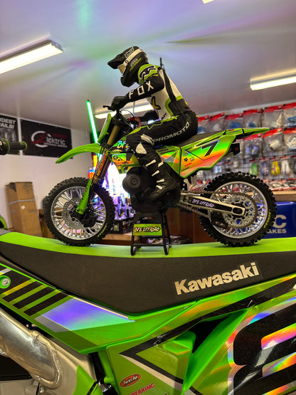 A green and black Kawasaki dirt bike displayed indoors on a stand, with a rider in black and white motocross gear leaning over it. The bike has visible Kawasaki branding and colorful decals.