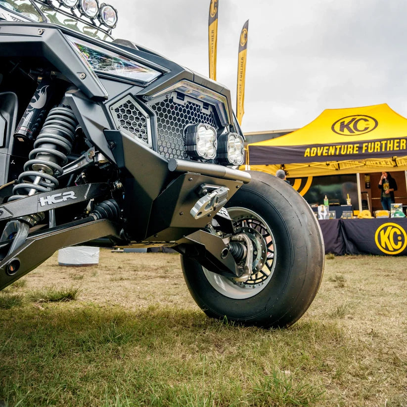 Close-up of a vehicle's front wheel and suspension system with a KC HiLiTES tent in the background.