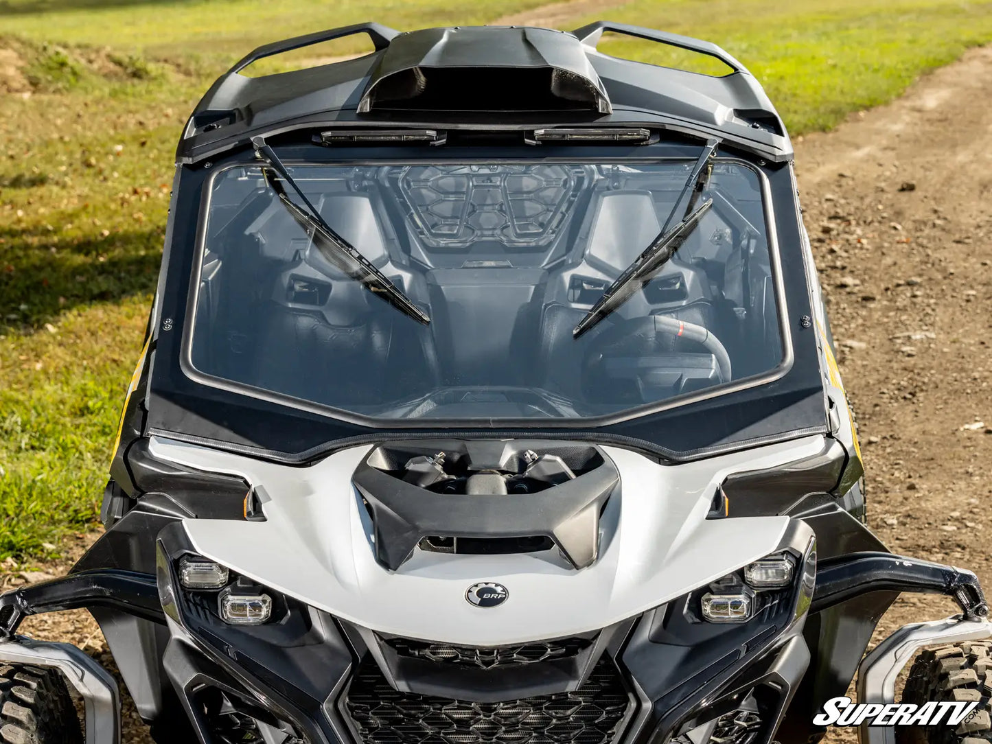 Front view of a Can-Am vehicle with a clear windshield and wipers on a grassy and dirt path background.