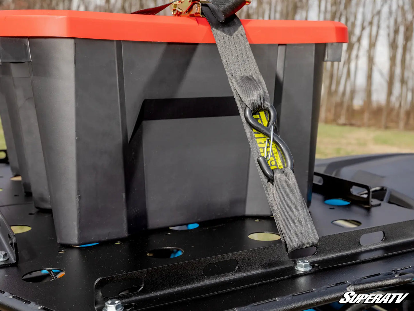 Gray storage box with a red lid and strap on a SuperATV vehicle, with trees in the background.