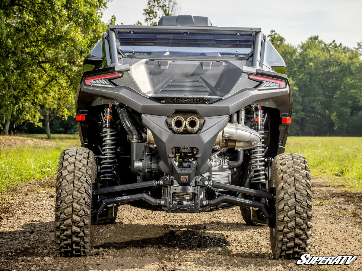 A rear view of a black Can-Am side by side vehicle with off-road tires, suspension, and exhaust system, parked on a dirt path with green trees in the background.