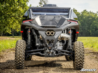 A rear view of a black Can-Am side by side vehicle with off-road tires, suspension, and exhaust system, parked on a dirt path with green trees in the background.