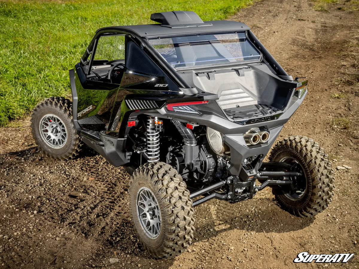 A black and gray Can-Am X10 side-by-side dune buggy with large off-road tires, shown from a rear three-quarter angle on a dirt track with grass in the background.
