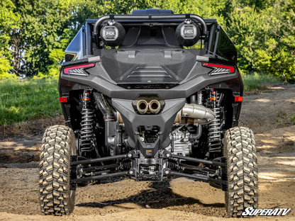 Rear view of a black off-road side-by-side UTV with large knobby tires, dual rear exhausts, and two round auxiliary lights mounted on the rear crossbar. The vehicle is photographed outdoors on a dirt trail with trees in the background.