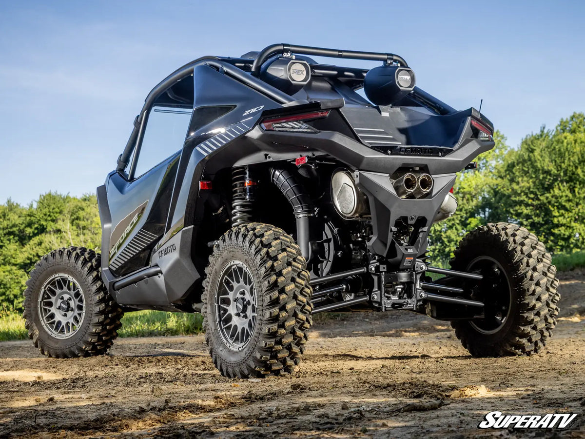 A black and gray Can-Am Maverick X3 off-road utility vehicle with large knobby tires, a roll cage, and auxiliary lights, parked on a dirt trail with trees and blue sky in the background.
