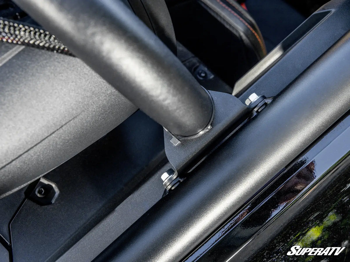 Close-up of a matte black roll bar installed over a car door frame, with visible mounting hardware and part of the interior seat in the background. A SuperATV logo watermark appears in the bottom right corner.