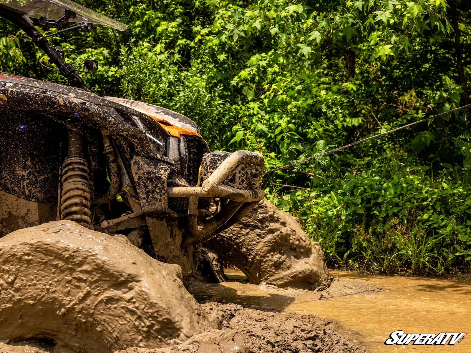 Mud-covered ATV on a rocky trail with lush greenery in the background, featuring the SuperATV logo.