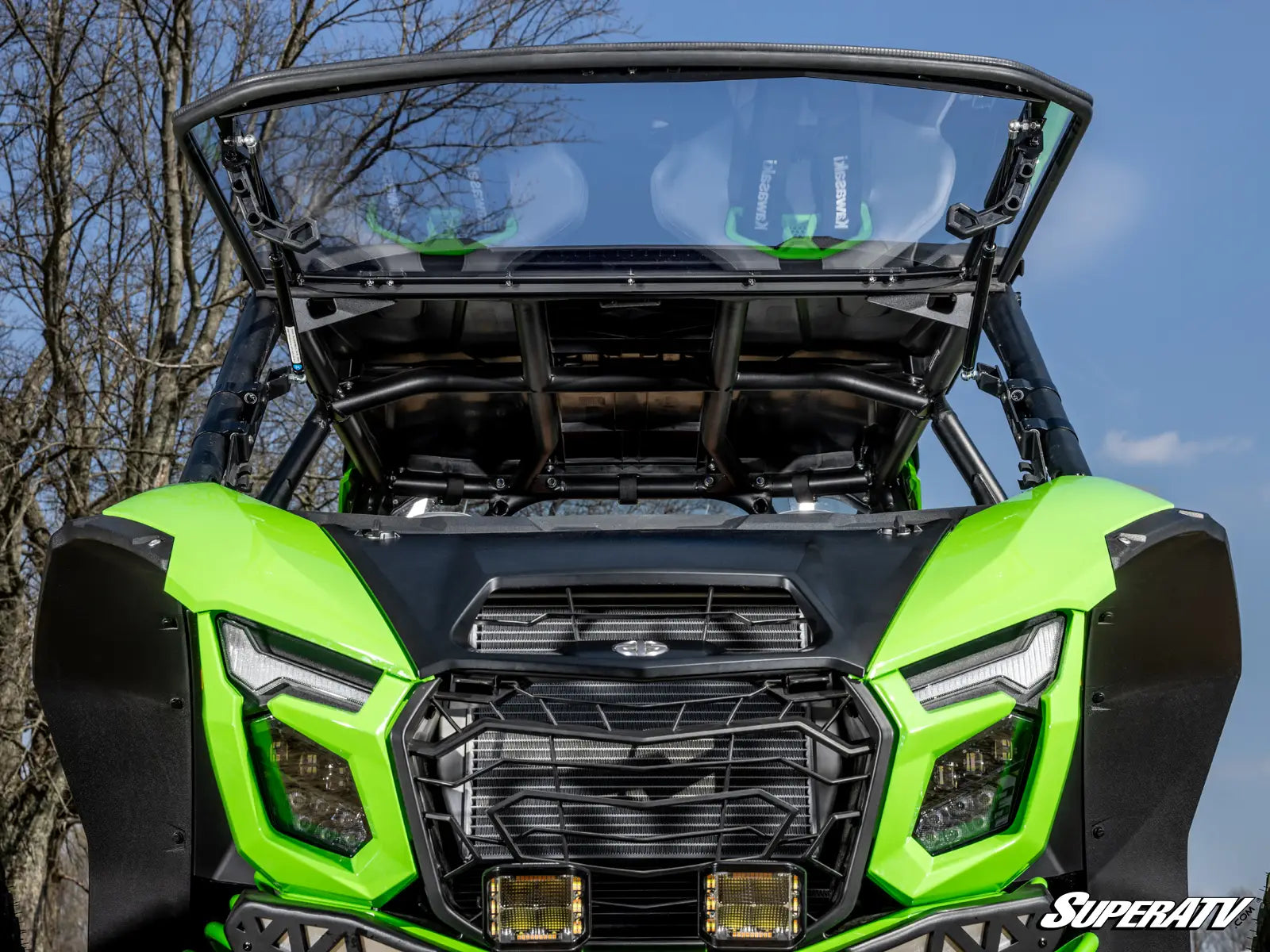 Green SuperATV vehicle with a clear windshield against a blue sky and tree background
