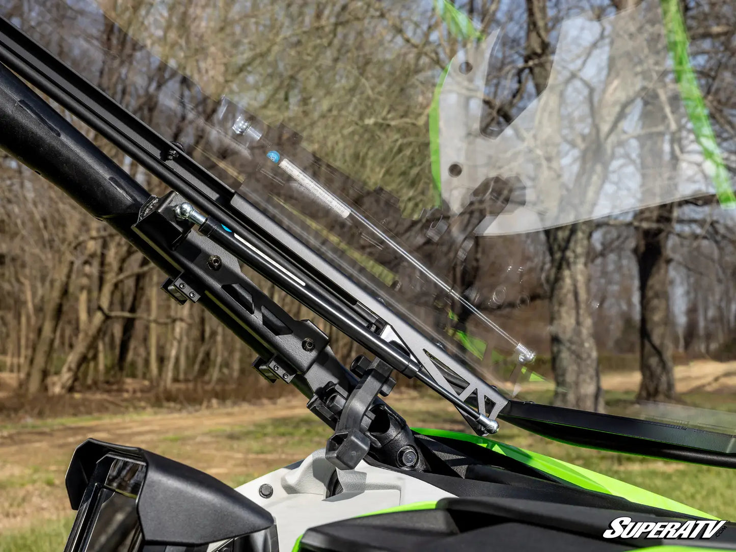 Close-up of a motorcycle windshield with a clear windshield washer arm and wiper arm assembly mounted on the frame, with a wooded outdoor background and a SuperATV logo in the corner.