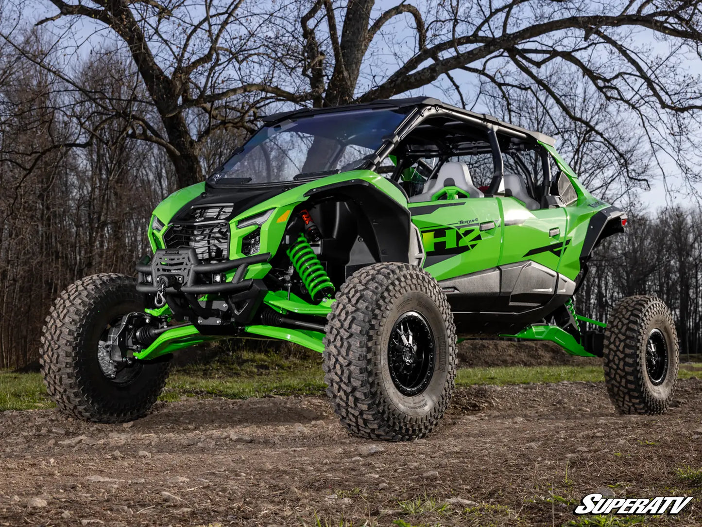 A bright green and black Can-Am Maverick X3 side-by-side off-road vehicle parked on a dirt trail with bare trees in the background. The vehicle features large knobby tires, a front bumper guard, and visible suspension components.