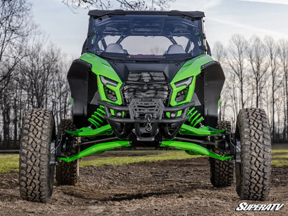 A front-facing view of a custom-built off-road vehicle with a black roll cage and body panels, featuring vibrant lime green accents on the fenders, bumpers, and suspension. The vehicle is equipped with massive, knobby off-road tires and is positioned on a dirt track with bare trees in the background.