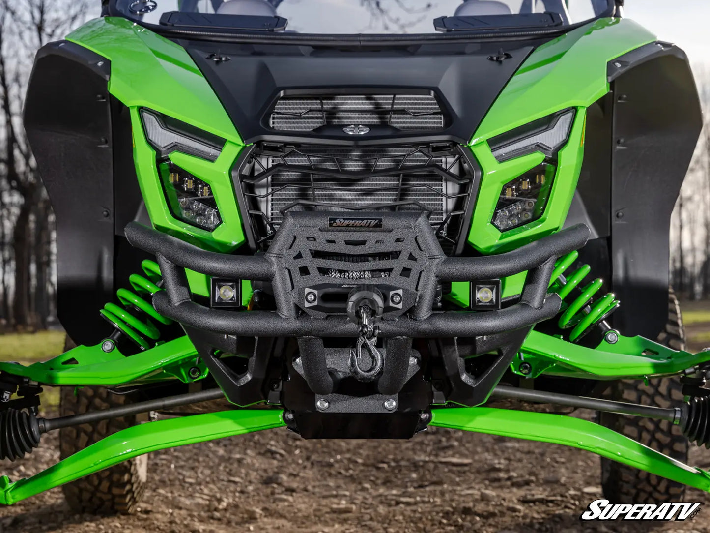A close-up front view of a bright green and black off-road side-by-side vehicle with a heavy-duty black front bumper and guard. The vehicle is outdoors on a dirt ground with trees in the background.