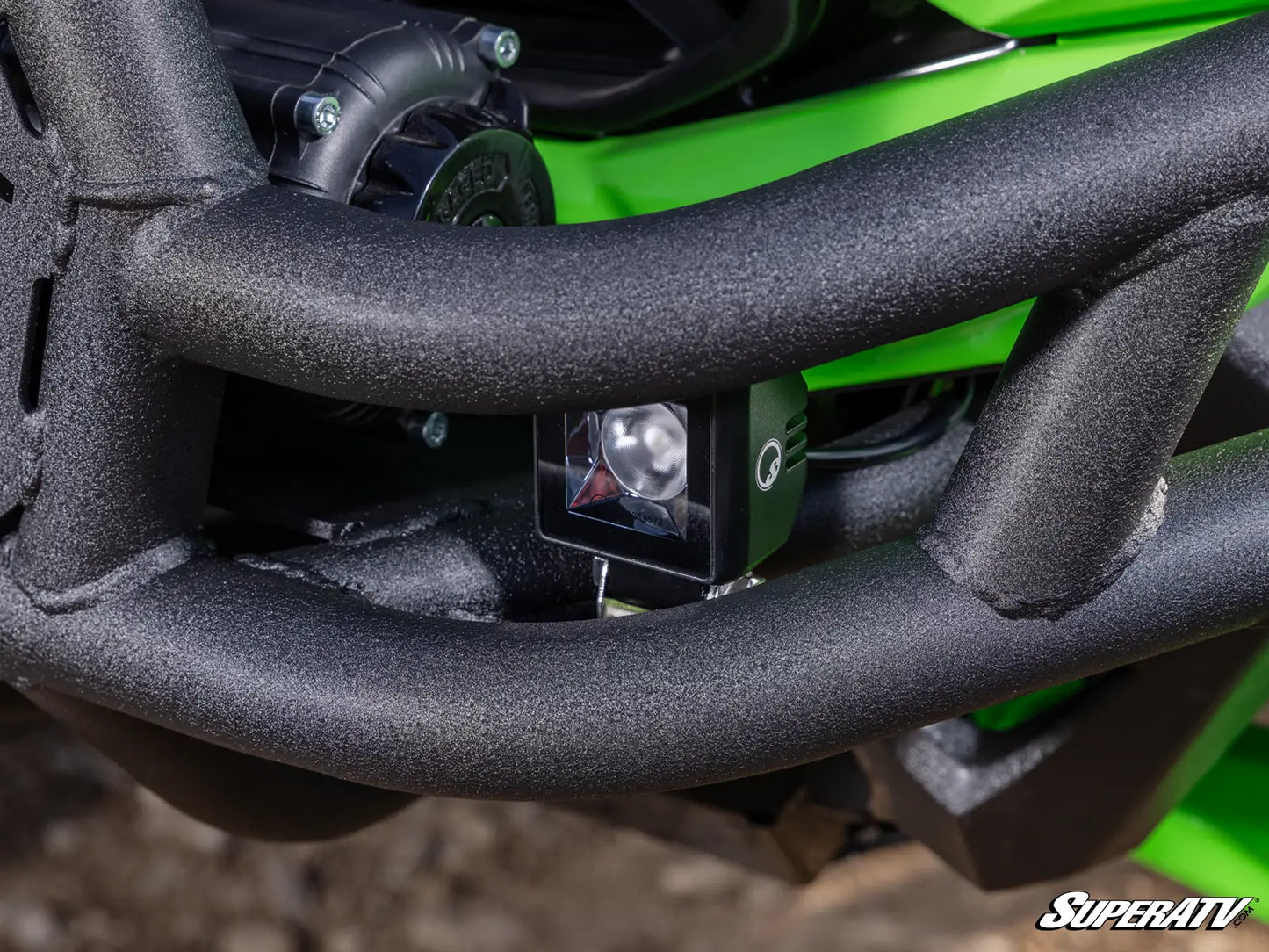 Close-up of a black off-road auxiliary LED light mounted on the front bumper of a green and black off-road vehicle, with a Superatv watermark in the bottom right corner.
