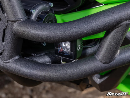 Close-up of a black off-road auxiliary LED light mounted on the front bumper of a green and black off-road vehicle, with a Superatv watermark in the bottom right corner.