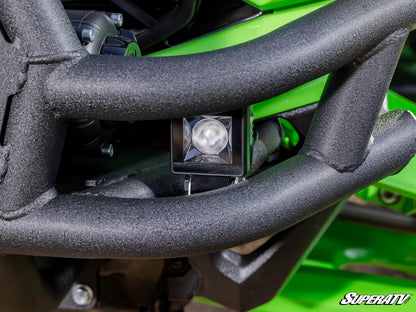 A close-up shot of a black square LED light mounted on a black tubular metal frame, set against a bright green vehicle body. A 'SUPERATV' logo watermark is visible in the bottom right corner.