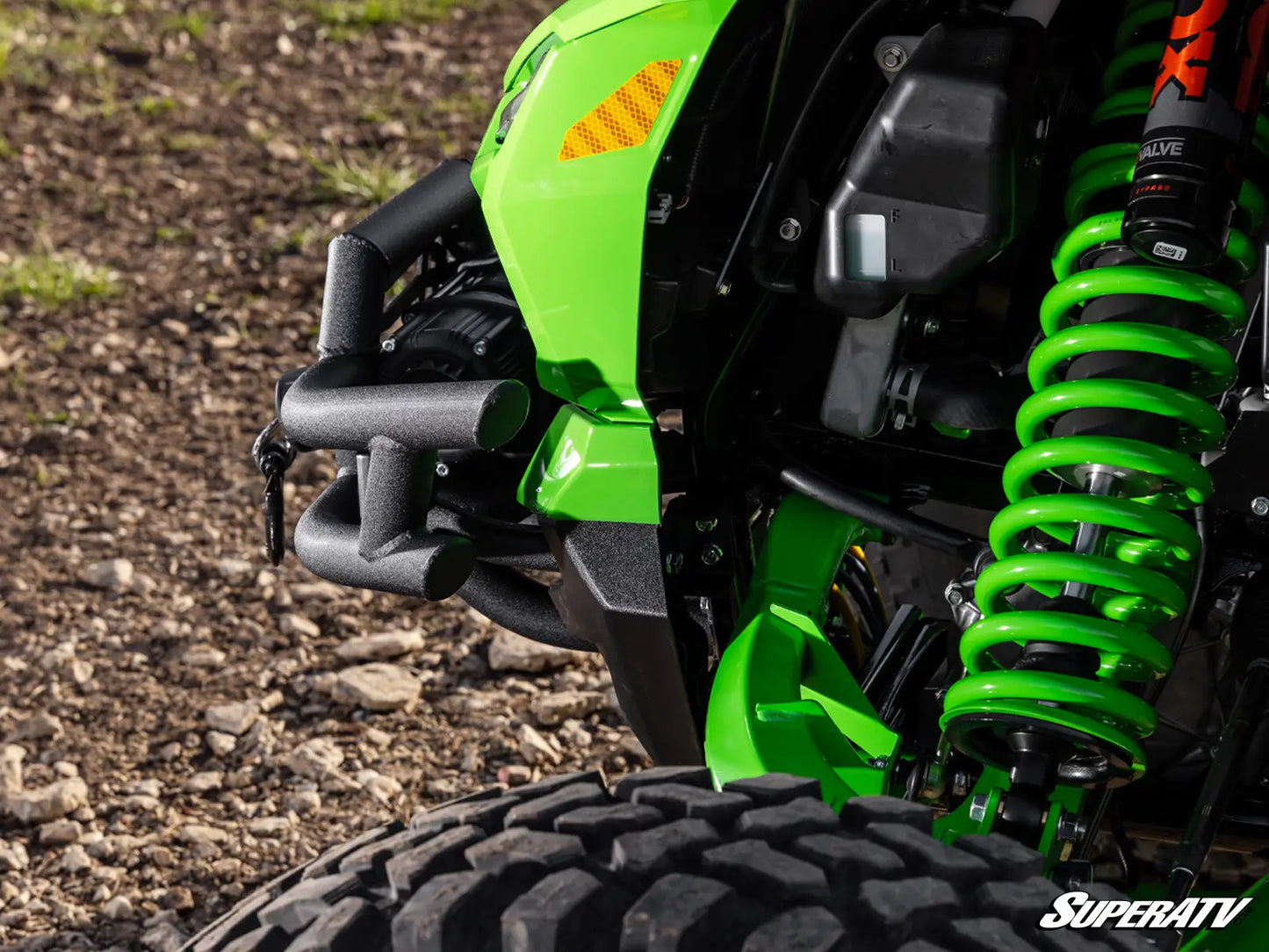 Close-up of a green and black off-road vehicle body panel with a bright green coil spring, black suspension components, and a tire on a gravel surface. A SuperATV logo watermark appears in the bottom right corner.