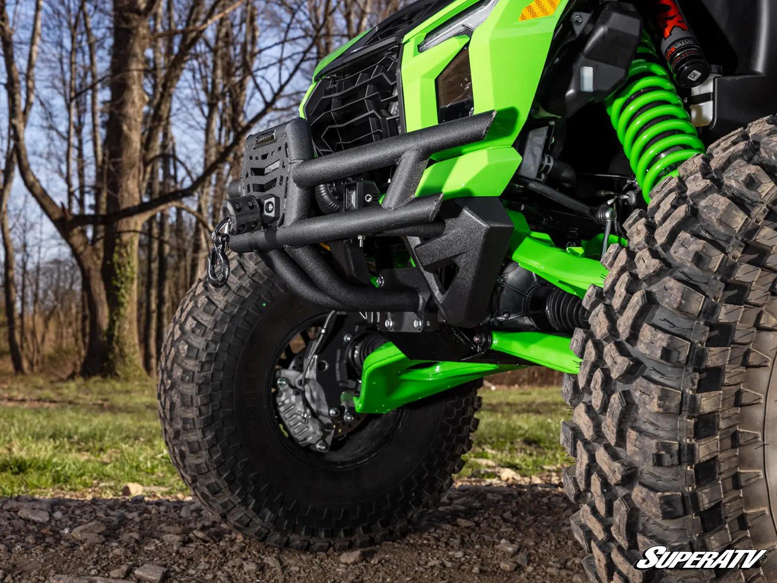 Close-up of a green and black off-road vehicle with knobby tires, a black front bumper guard, and bright green suspension springs, photographed outdoors on a dirt path with trees in the background.