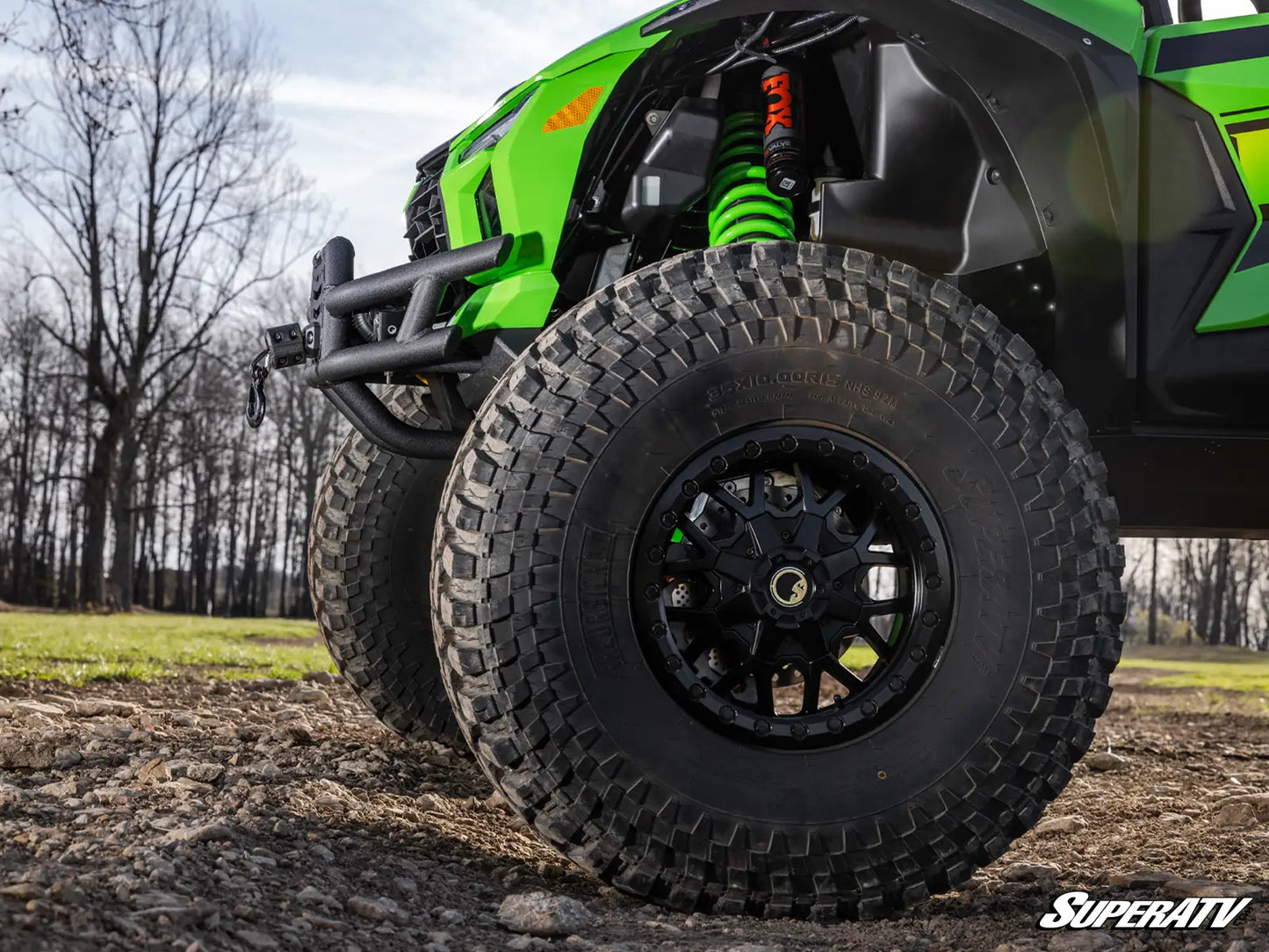 Close-up of a green and black off-road vehicle wheel with a black all-terrain tire, suspension, and forest background.