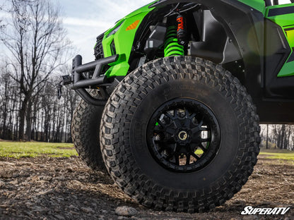 Close-up of a green and black off-road vehicle wheel with a black all-terrain tire, suspension, and forest background.