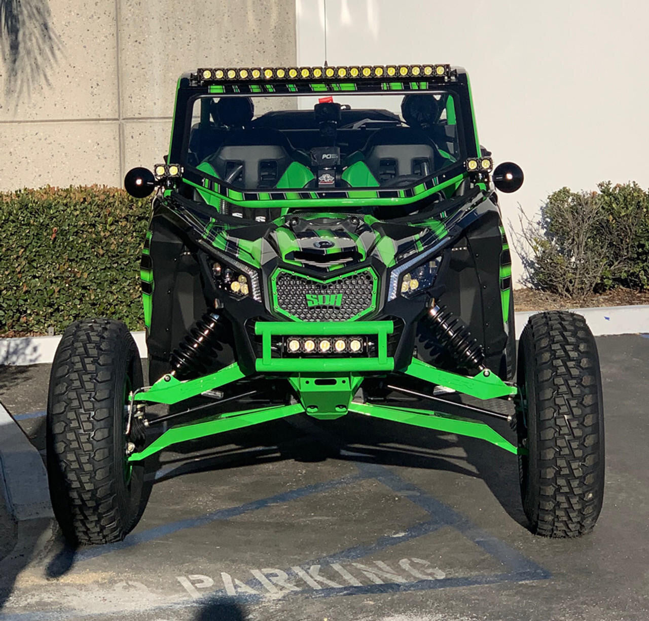 A custom-built off-road side-by-side UTV with a striking black and neon green color scheme, parked in a paved parking lot. The vehicle features a roll cage, a front bumper with integrated LED light bars, and oversized knobby tires.