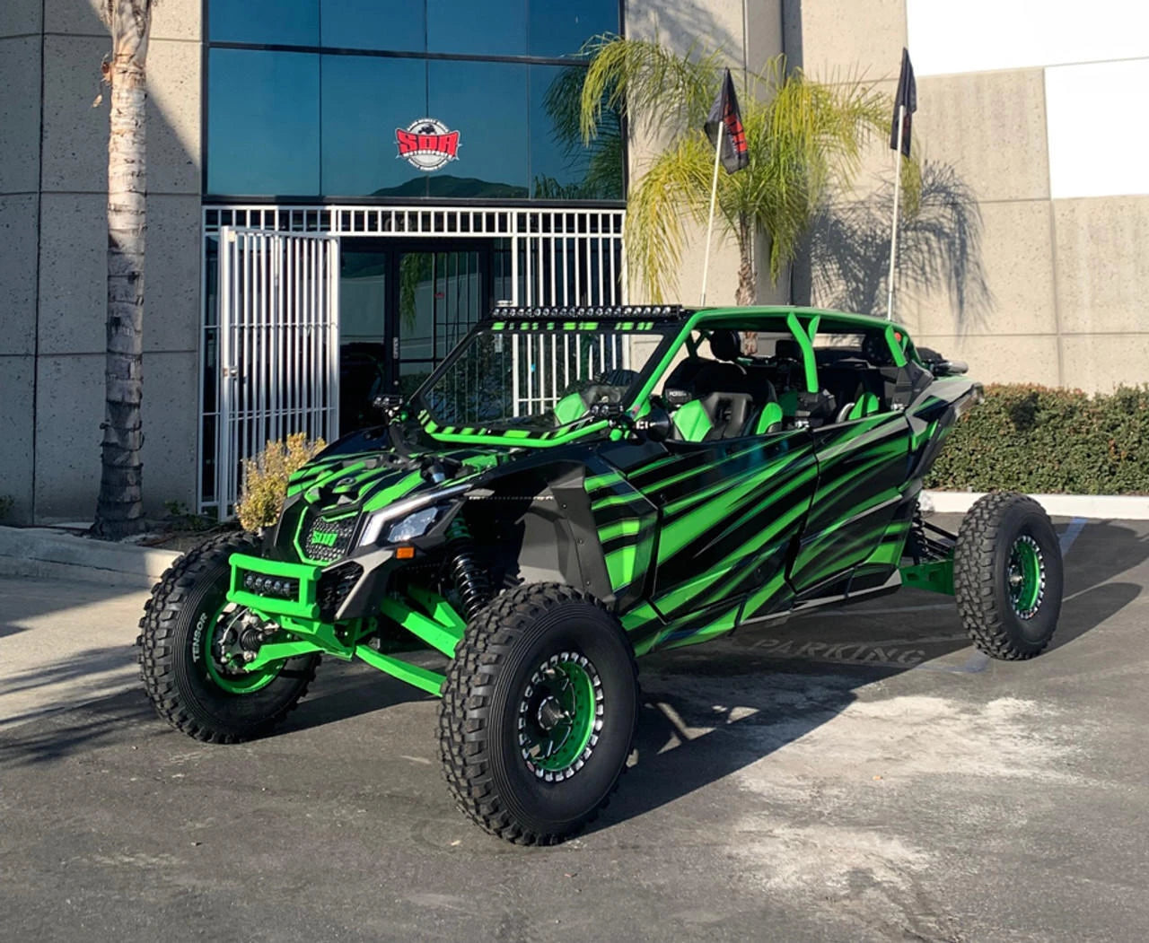 A green and black Can-Am X-Series side-by-side off-road vehicle parked on asphalt in front of a modern building with palm trees.