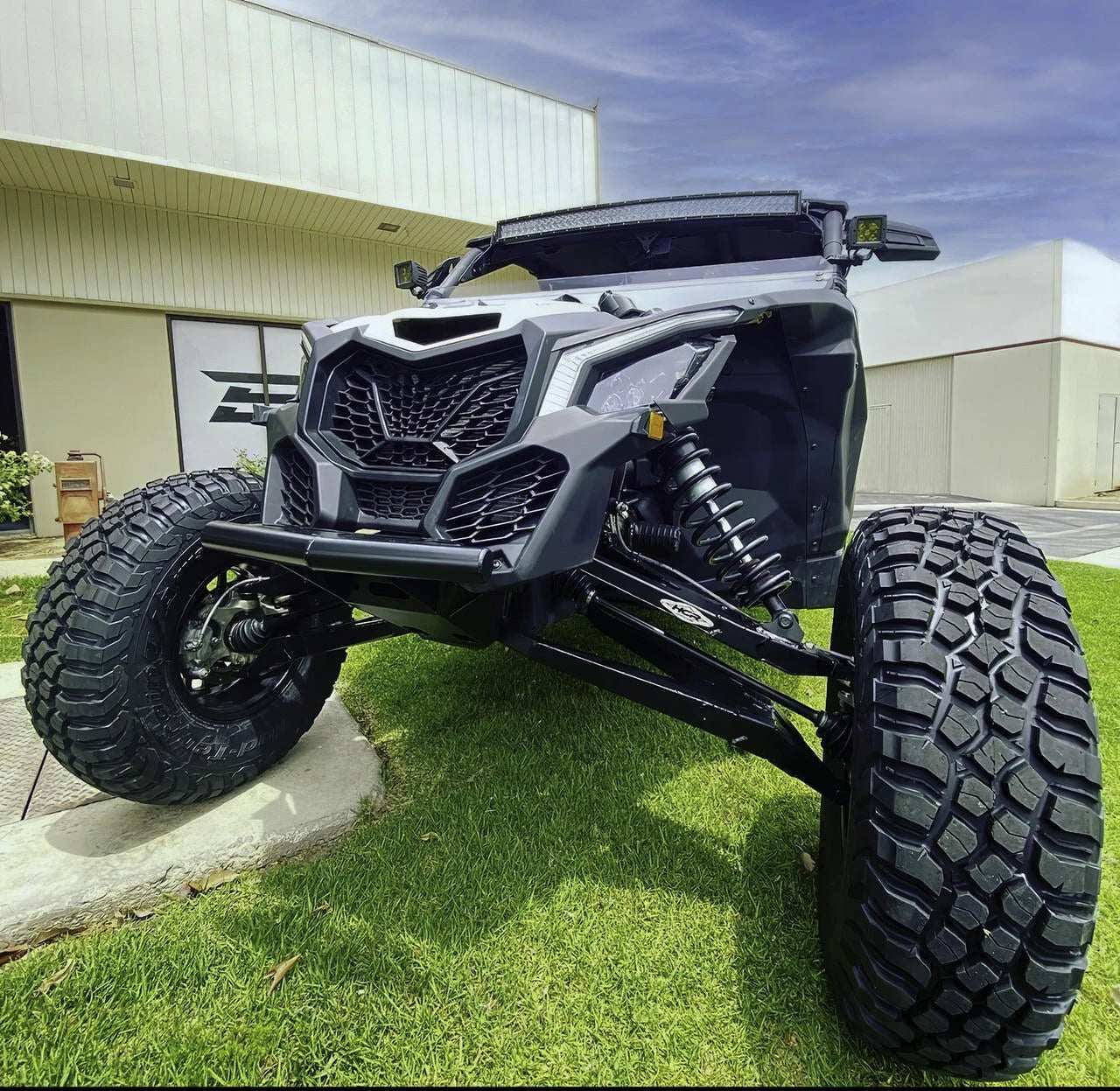 A black Can-Am side-by-side UTV with oversized knobby tires, a front bull bar, and a custom roll cage, parked on a grassy lawn in front of a light-colored commercial building.