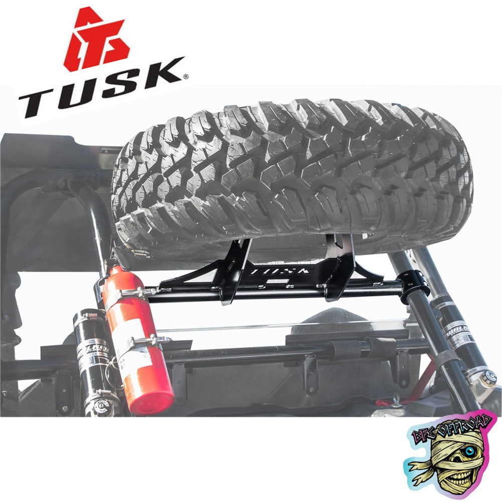 Tusk brand spare tire carrier rack mounted on an off-road vehicle frame, holding a large off-road tire. A red fire extinguisher is attached to the frame.